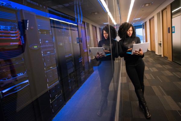 woman looking into her computer and leaning against a glass wall. behind the glass wall is computer boards glowing a somber blue color. 