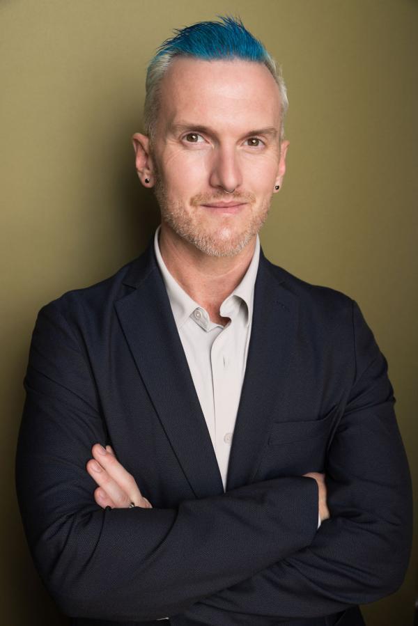 Bryan Stanton, average height, slightly chubby, white person, with blonde hair and blue streaks standing with his arms crossed in a grey shirt and blue jacket.