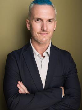 Bryan Stanton, average height, slightly chubby, white person, with blonde hair and blue streaks standing with his arms crossed in a grey shirt and blue jacket.